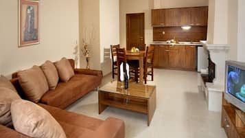 Ein Wohnzimmer mit braunem Sofa, einem Glastisch und einem Essbereich mit Holzkstühlen und einem kleinen Tisch. Im Hintergrund ist eine Küche mit Holzschränken zu sehen.