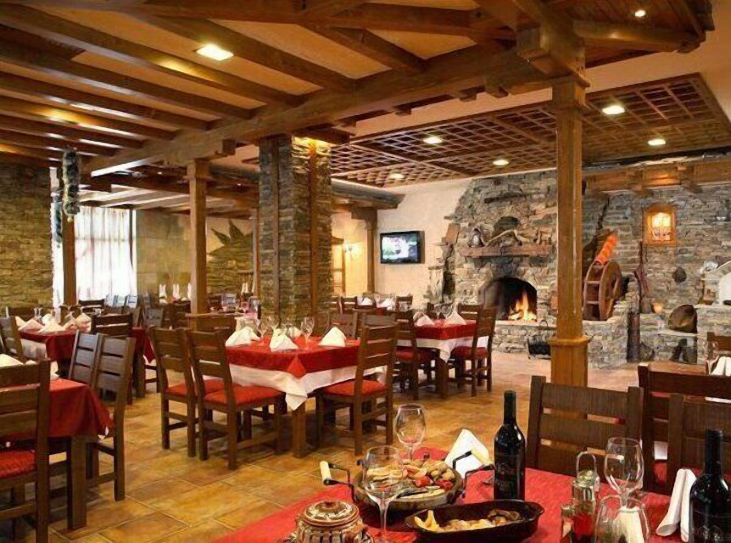 Ein Restaurant mit Holzbalken, Steinwänden und einem Kamin. Die Tische sind mit roten Tischdecken und Weingläsern gedeckt.