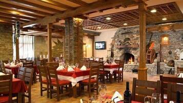 Ein Restaurant mit Holzbalken, Steinwänden und einem Kamin. Die Tische sind mit roten Tischdecken und Weingläsern gedeckt.