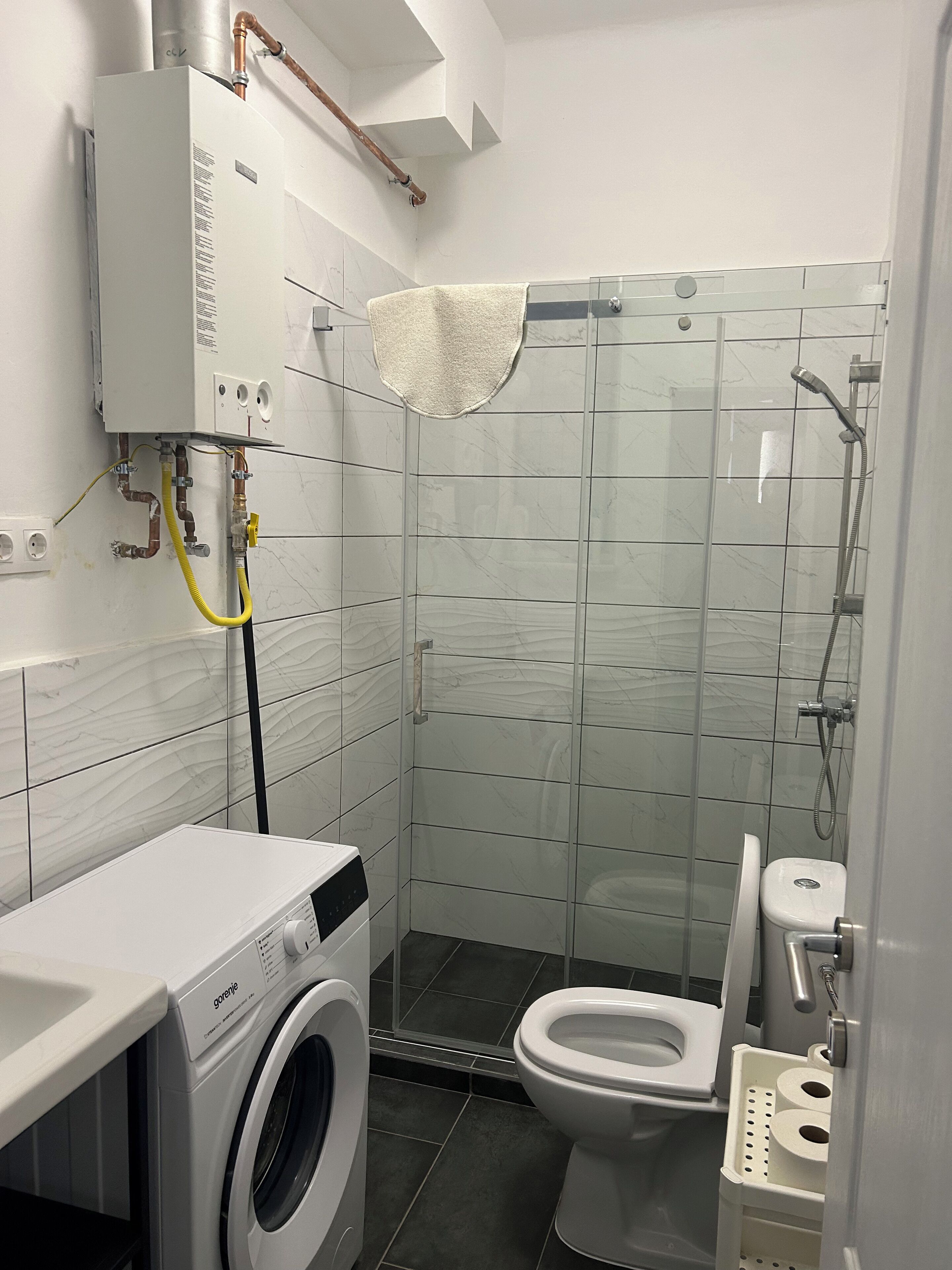 A compact bathroom with a washing machine, toilet, and a glass shower enclosure.