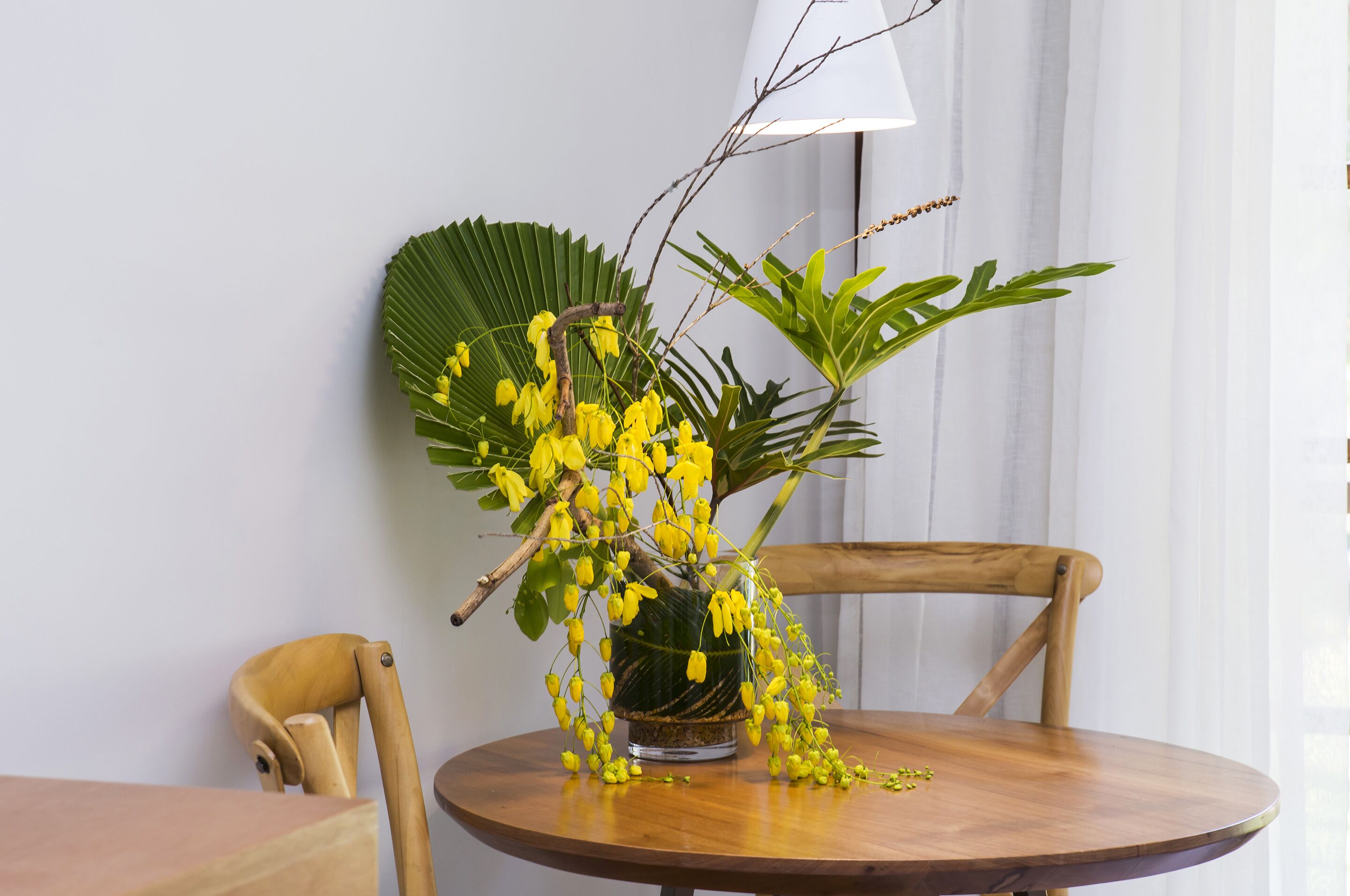 A round wooden table with a clear glass vase containing yellow flowers and green foliage.