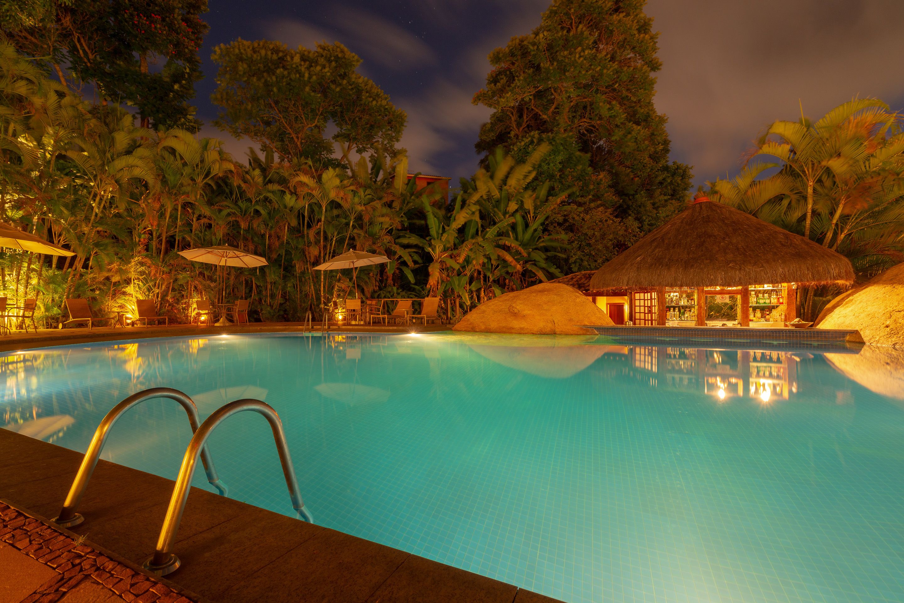 A swimming pool with a thatched-roof structure and palm trees in the background.