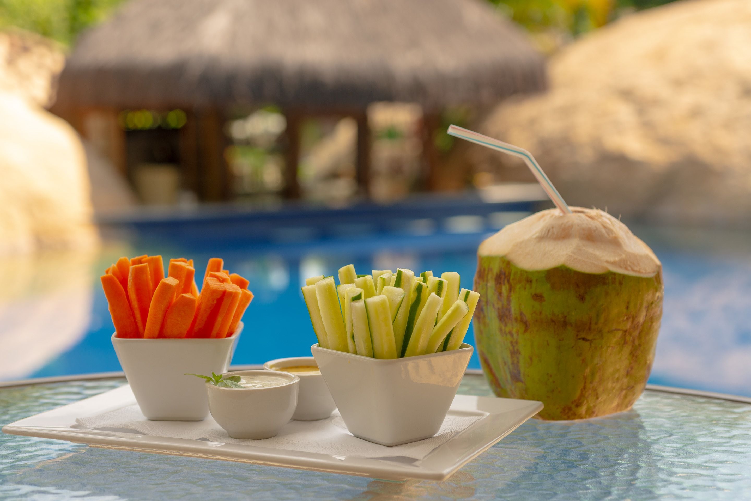 A tray with two bowls of vegetable sticks, a coconut drink, and two small bowls of dip, set against a poolside view.