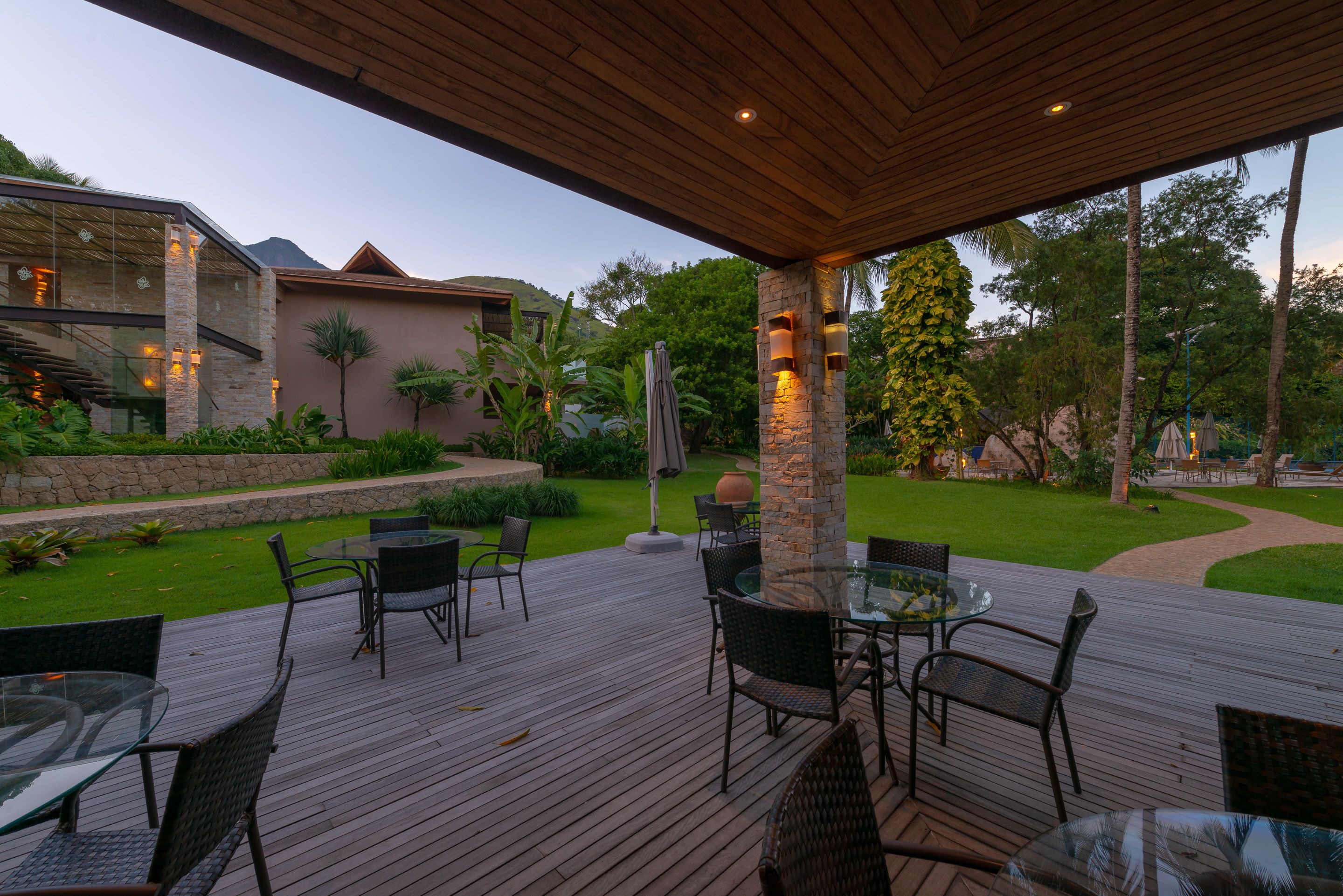 A covered outdoor patio with glass tables and chairs, surrounded by a landscaped garden with a stone wall and palm trees.