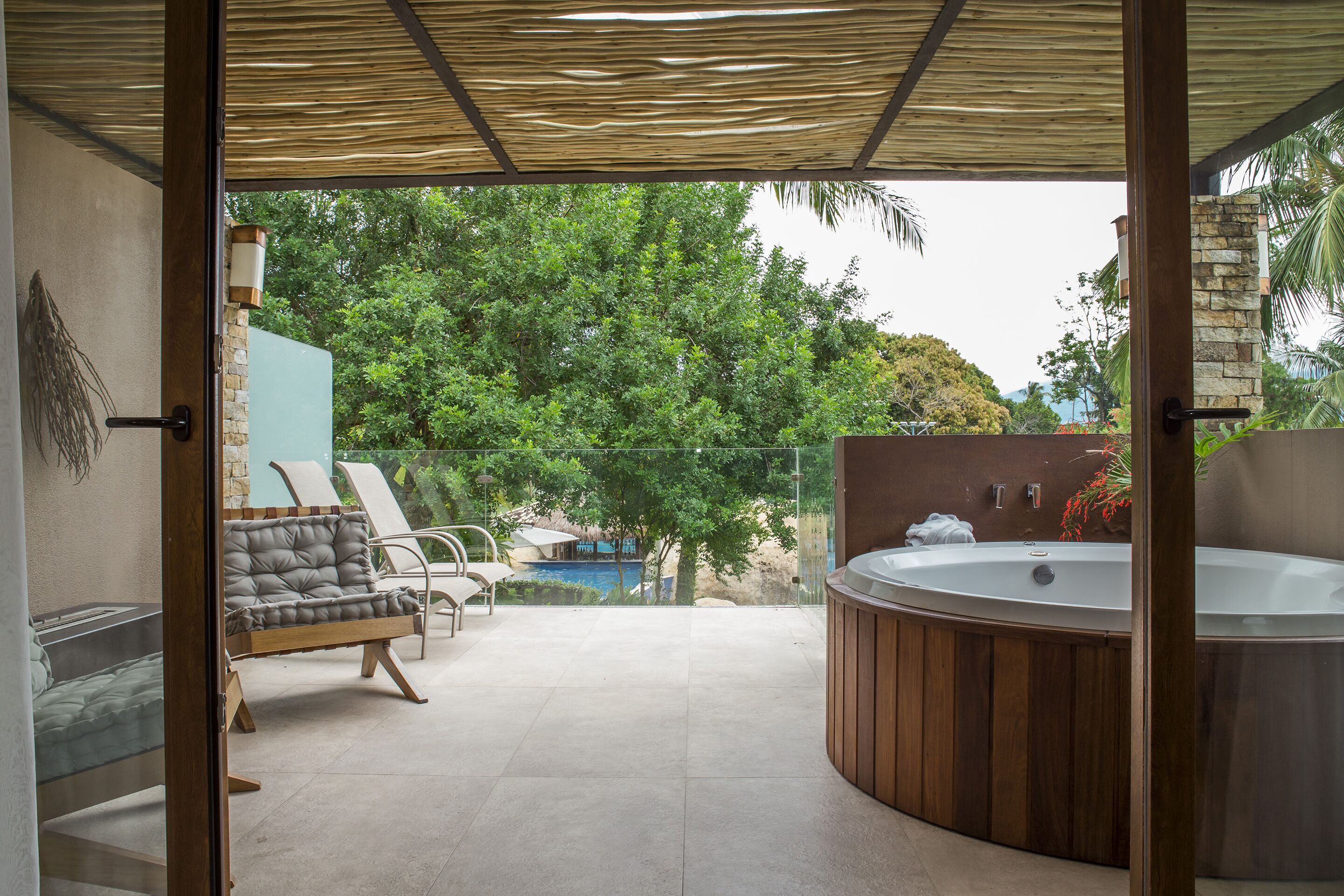A modern outdoor patio with a hot tub, lounge chairs, and a view of a pool and trees.