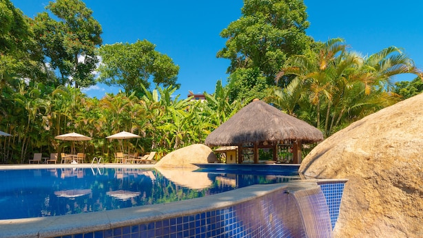 Een zwembad met helderblauw water, omringd door rotsen en palmbomen, met ligbedden en parasols.
