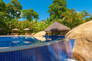Una piscina con agua azul cristalina, rodeada de rocas y palmeras, con sillas de descanso y sombrillas.
