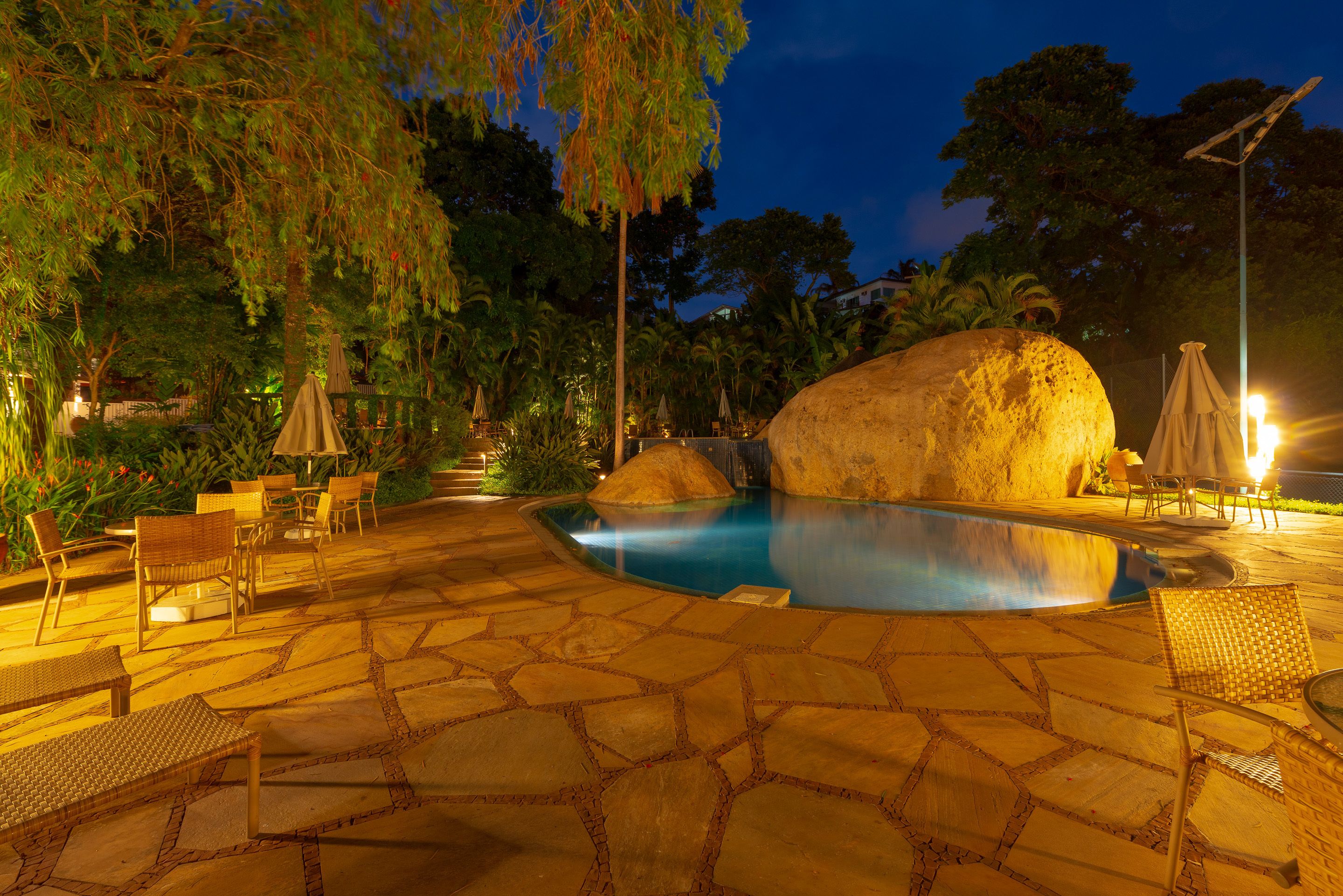 Un área de piscina exterior de noche con suelo de piedra, sillones reclinables y sombrillas.