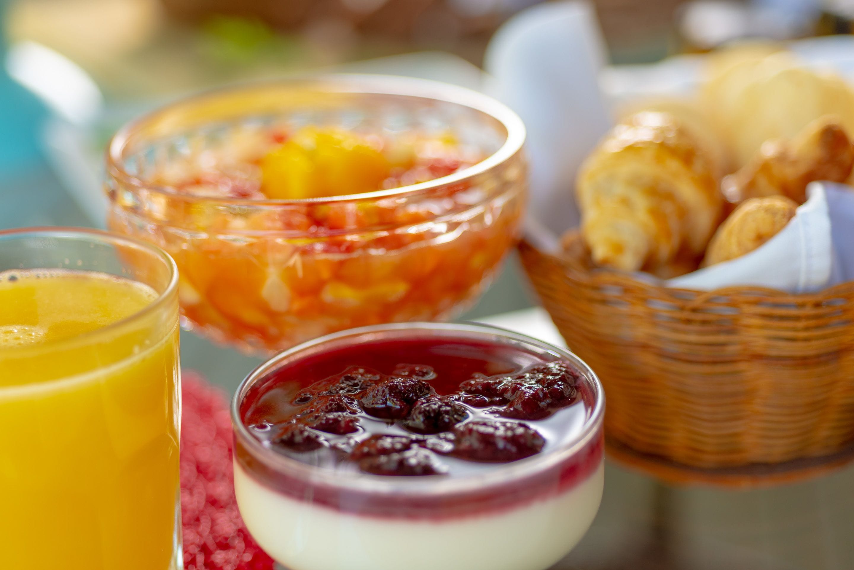 Una mesa de desayuno con un vaso de jugo de naranja, un bol de ensalada de frutas, un postre con bayas y una cesta de croissants.