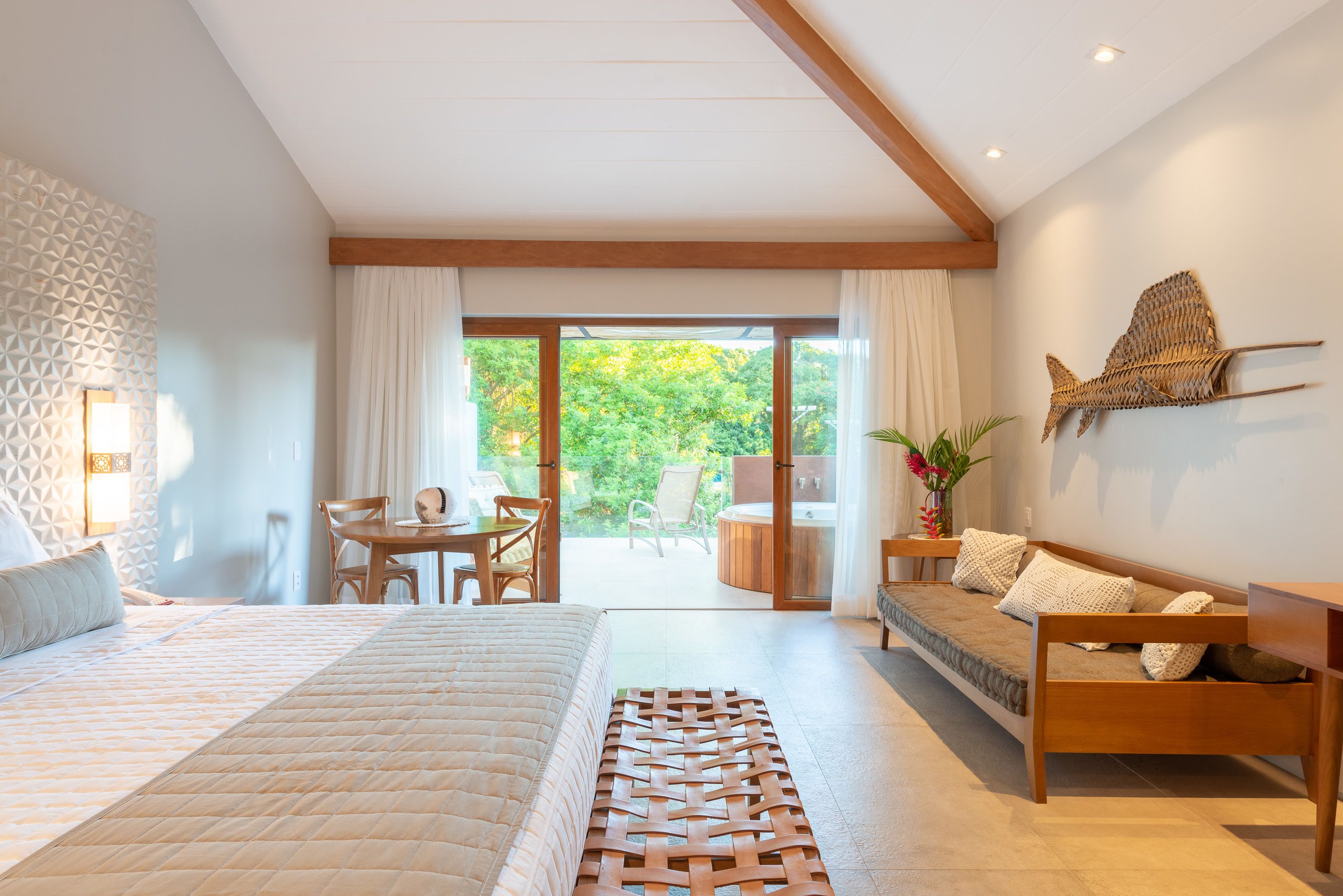 A modern hotel room with a large bed, a wooden bench, a dining area, and a view of the garden through the sliding doors.