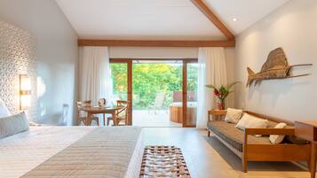 A modern hotel room with a large bed, a wooden bench, a dining area, and a view of the garden through the sliding doors.