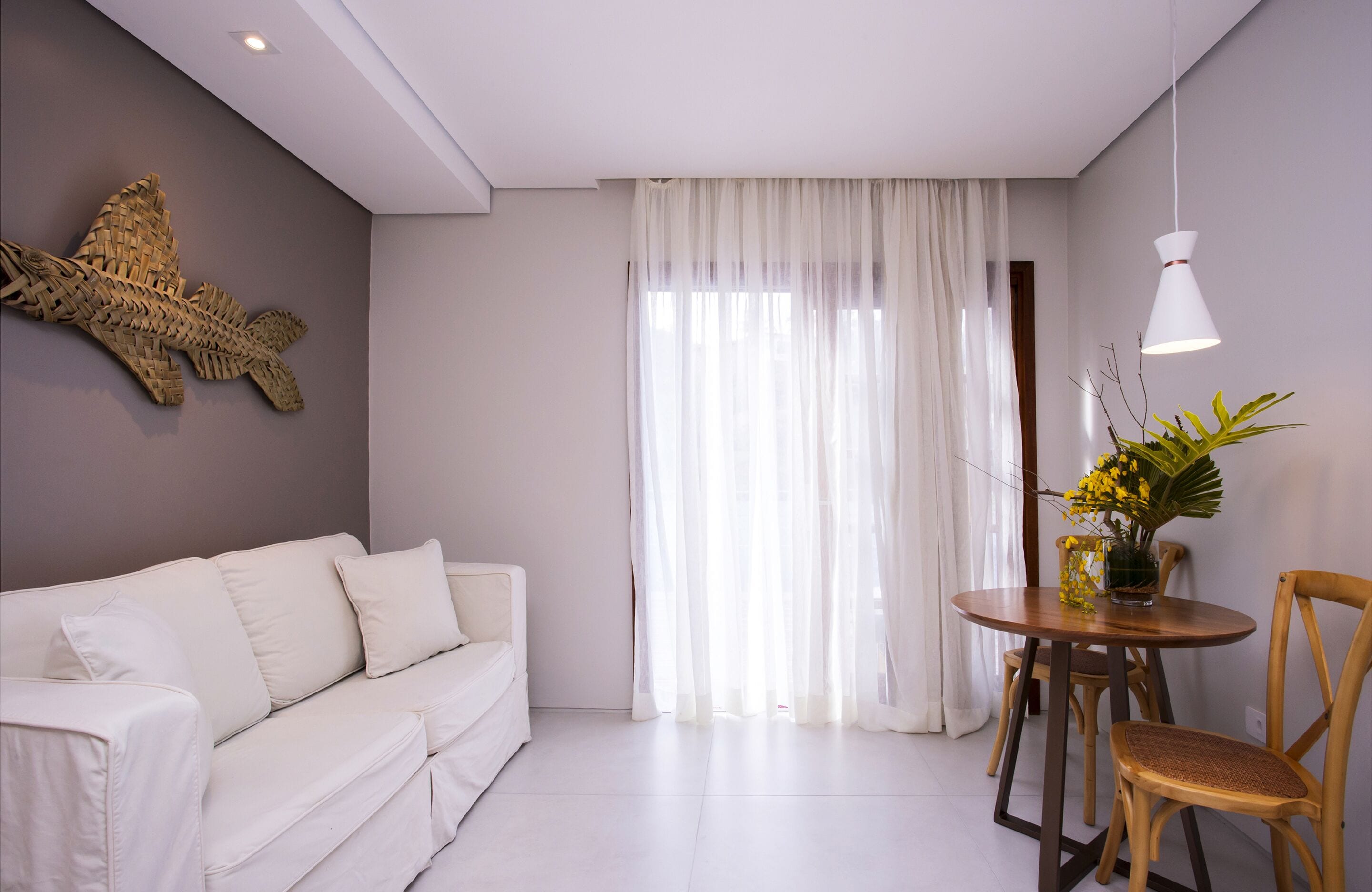 A modern living room with a white sofa, a round wooden dining table with two chairs, a decorative fish wall art, sheer white curtains, and a hanging white pendant light.