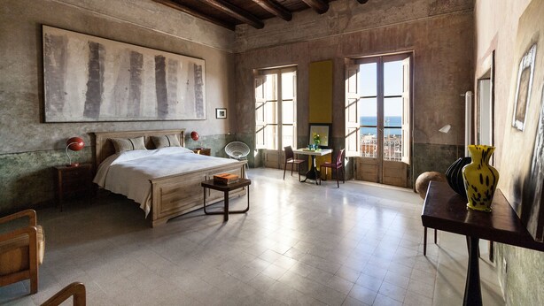 Une chambre spacieuse avec un grand lit, un bureau et une vue sur la mer par de grandes fenêtres ouvertes.