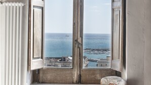 A room with a bay window overlooking the sea, a chair with a cushion, and a wooden dresser.