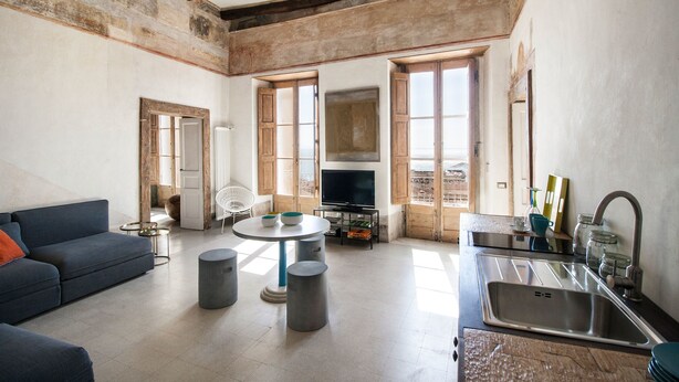 A spacious living room with a sofa, a round table, and a television. There is a kitchen area with a sink and cabinets.