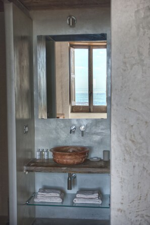 A bathroom with a large mirror, a wooden cabinet, a glass shelf with towels, and a view of the sea through a window.
