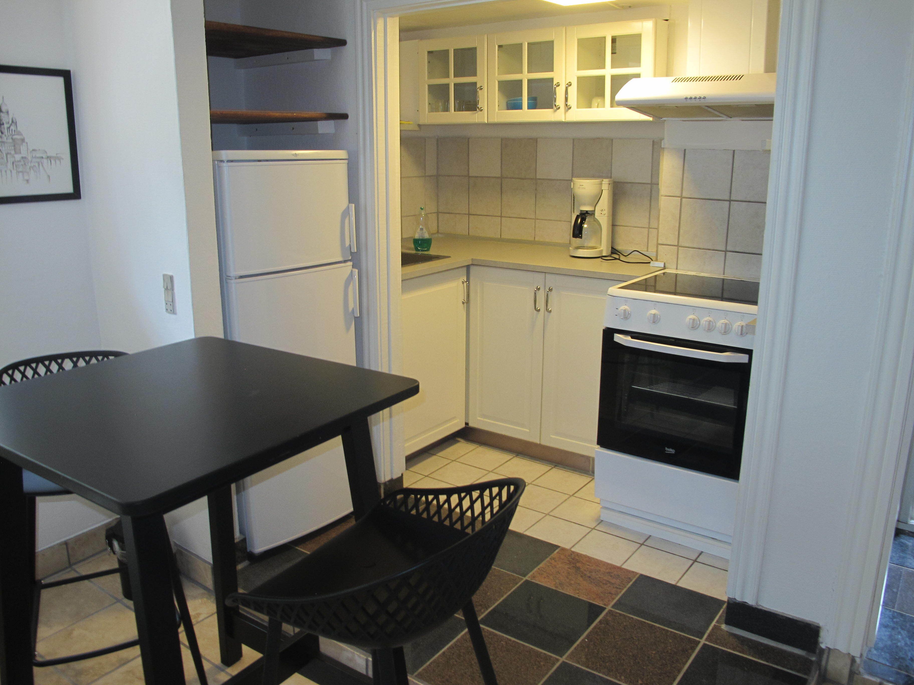 Una cocina compacta con armarios blancos, encimera negra, refrigerador blanco, estufa y una silla negra.