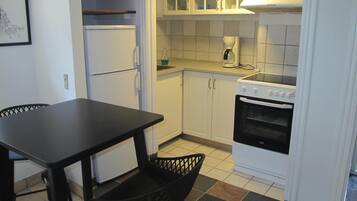 Una cocina compacta con armarios blancos, encimera negra, refrigerador blanco, estufa y una silla negra.
