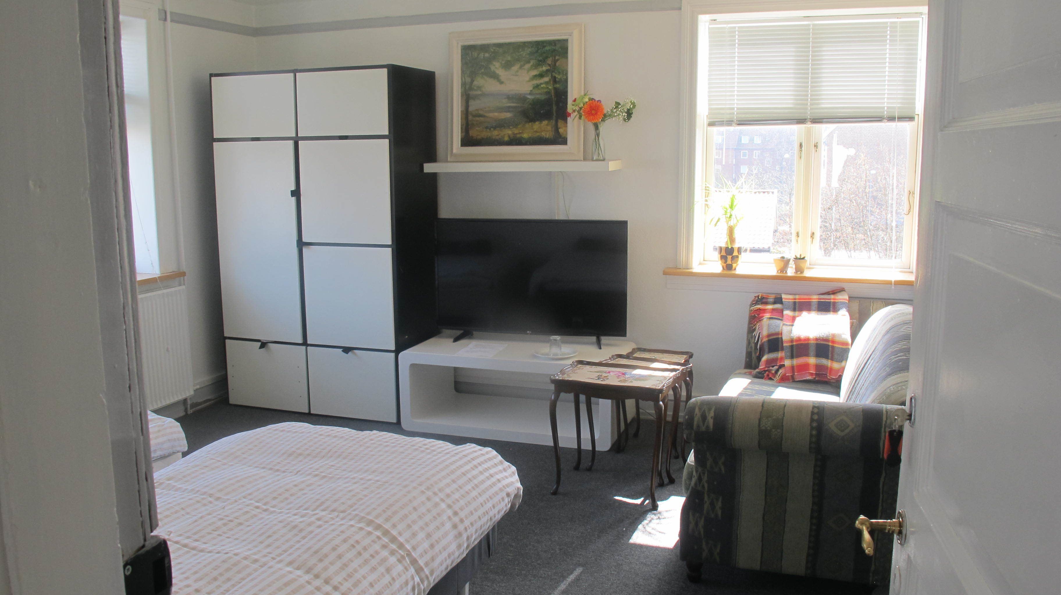 Een kleine kamer met een bed, een tafeltje, een televisie, een kledingkast en een raam met jaloezieën.