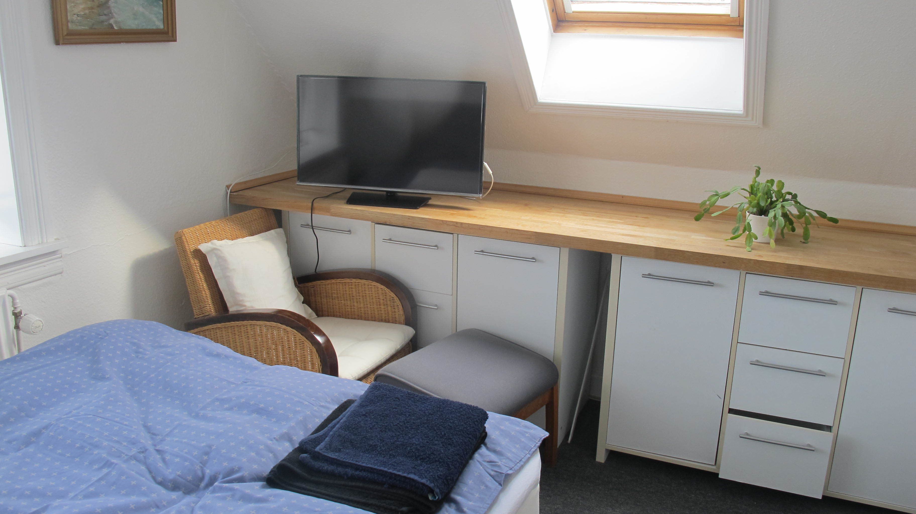 Quarto com cama, poltrona, escrivaninha e televisão, além de uma janela.