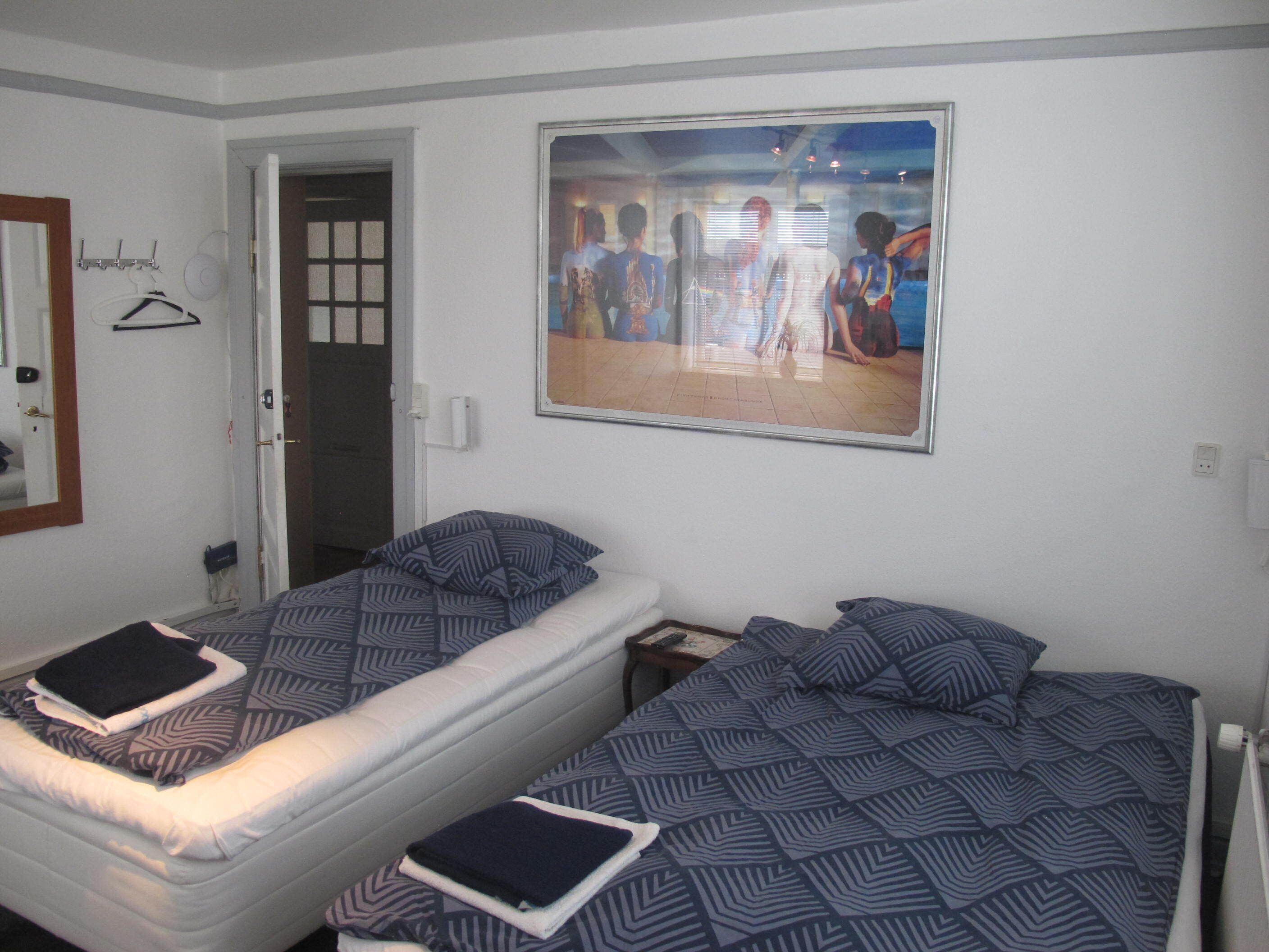 Une chambre avec deux lits simples, une porte, un porte-manteau et un tableau encadré au mur.