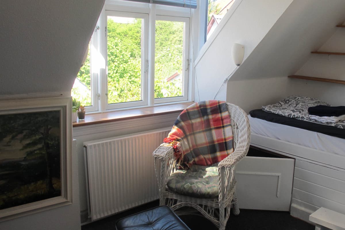 Ein Schlafzimmer mit schrägen Wänden, einem Fenster mit Blick auf Grünflächen, einem Korbsessel, einem Bett mit gemusterten Bettbezügen und einer Heizung.
