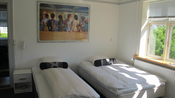 A room with two single beds, a painting on the wall, a window with blinds, and a radiator.