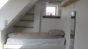 A small, single-bed room with a sloped ceiling, a window, a mirror, and a wooden shelf.