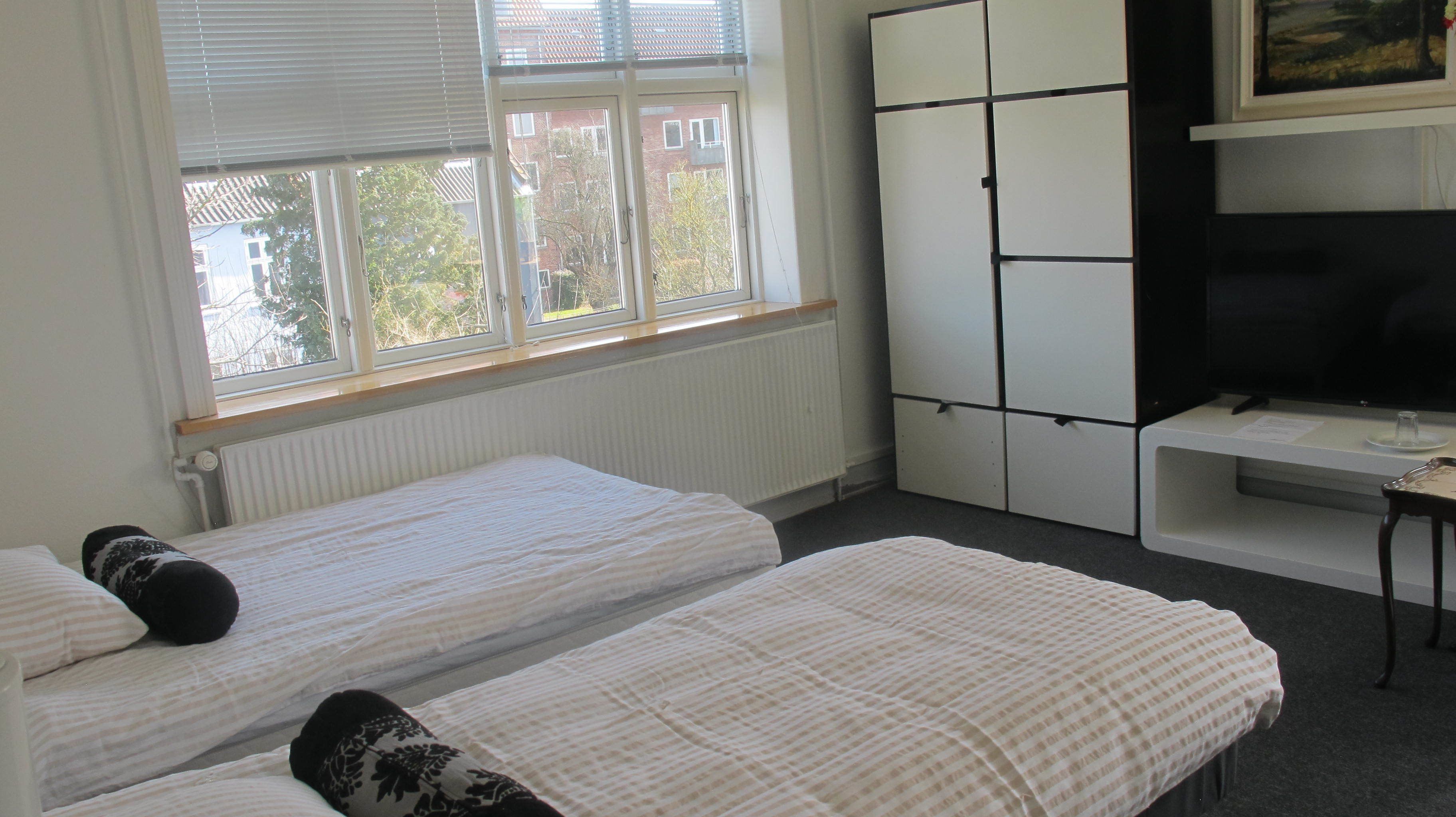 Een kamer met twee éénpersoonsbedden, een radiator, een raam met zonwering, een kast en een televisietoestel op een standaard.
