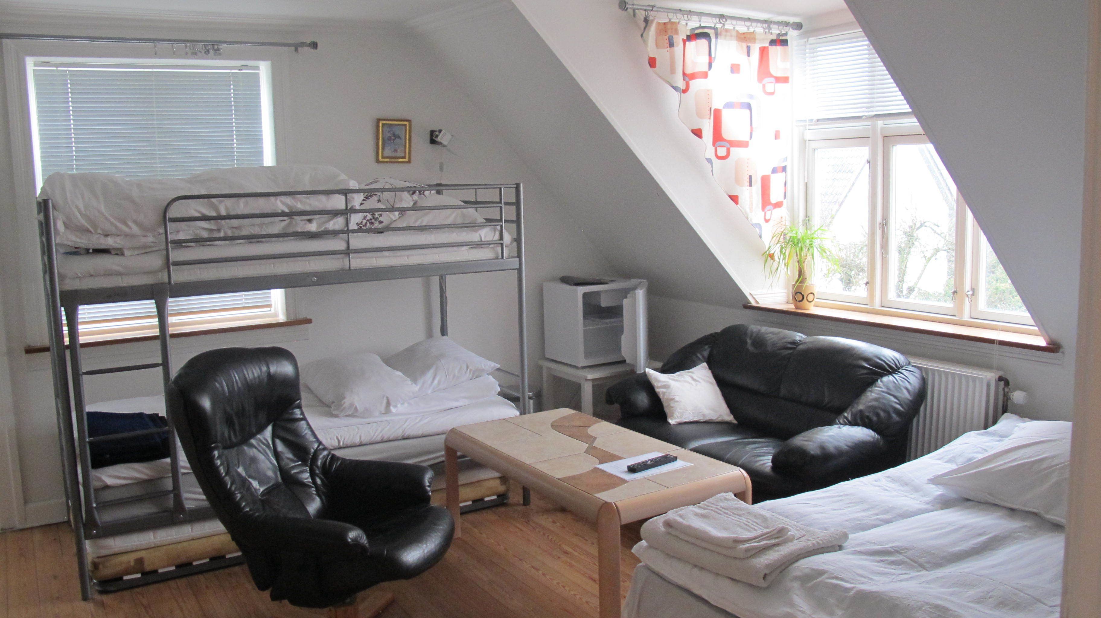 Ein Schlafsaalzimmer mit Etagenbetten, einem Ledersessel, einem kleinen Tisch und einem Fenster mit Blick auf Bäume.