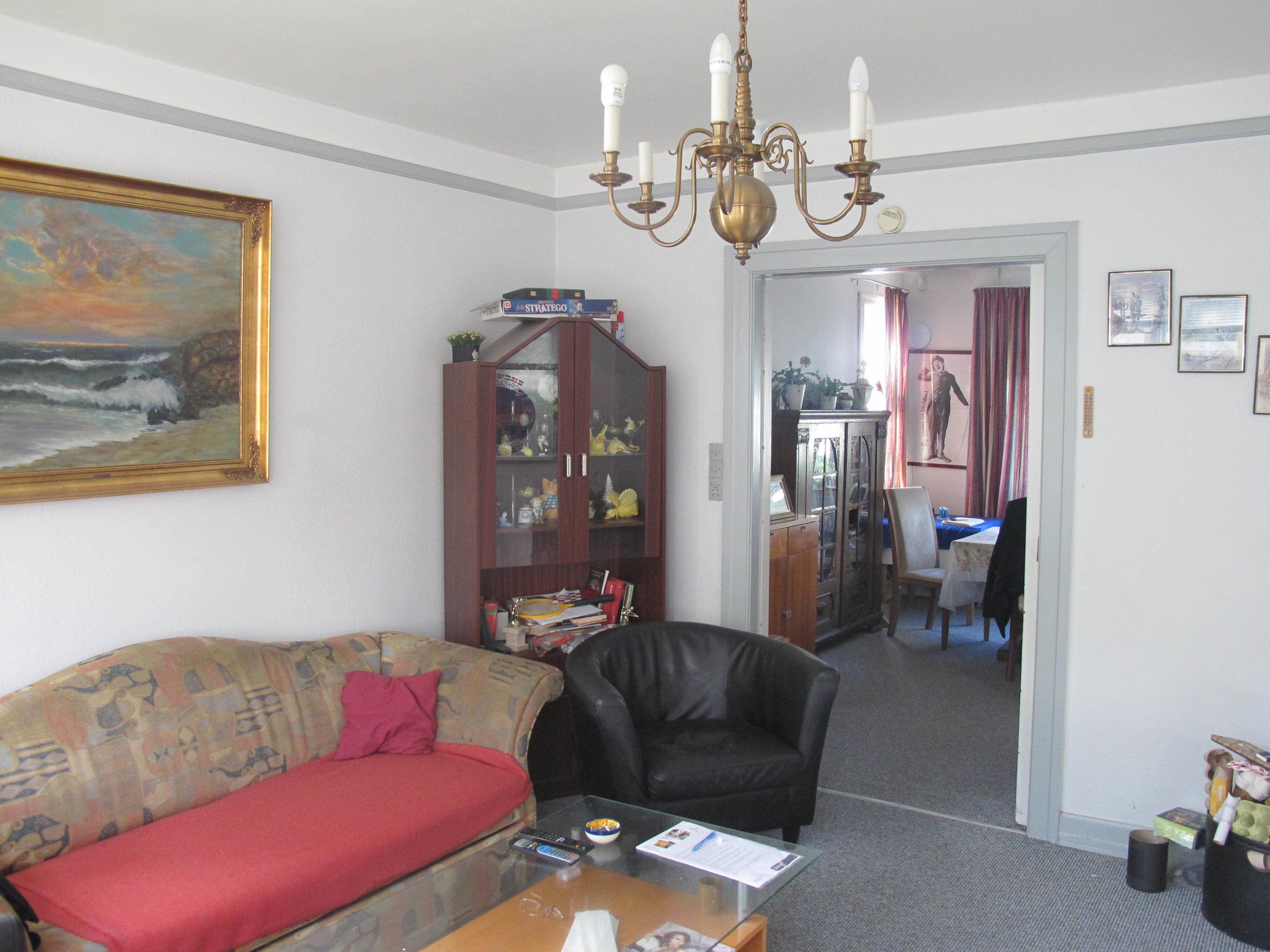 Ein Wohnzimmer mit einer Couch, einem schwarzen Ledersessel, einem Glastisch und einer Chandelier. Es gibt einen Schrank mit Glastüren und ein Bild an der Wand.