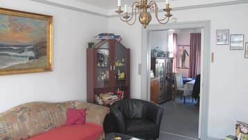 Ein Wohnzimmer mit einer Couch, einem schwarzen Ledersessel, einem Glastisch und einer Chandelier. Es gibt einen Schrank mit Glastüren und ein Bild an der Wand.