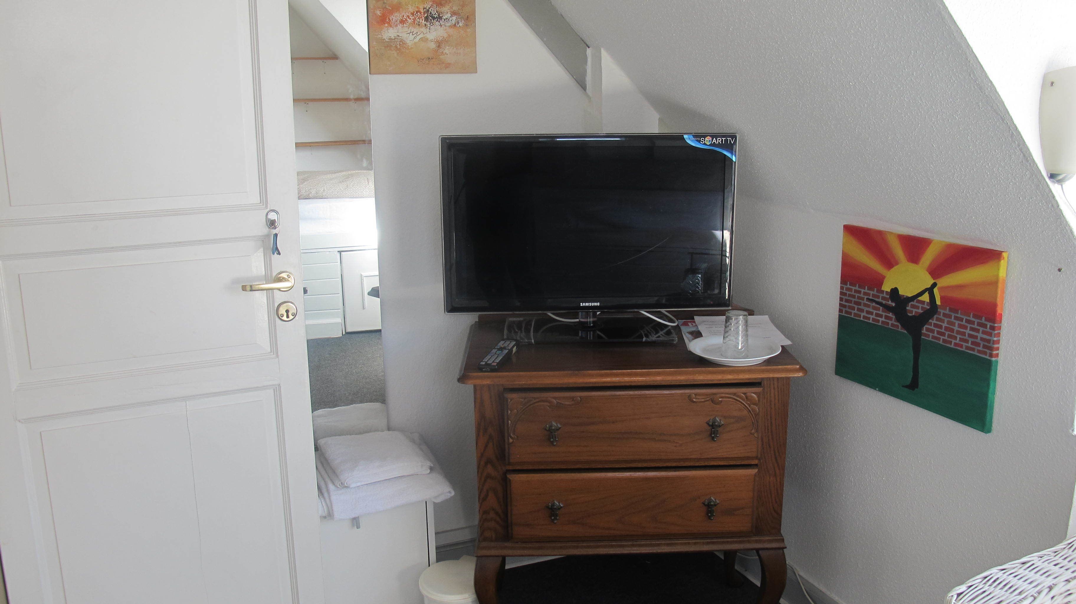 Une chambre à coucher avec une télévision posée sur une commode en bois, un tableau accroché au mur et une porte donnant sur une salle de bain.