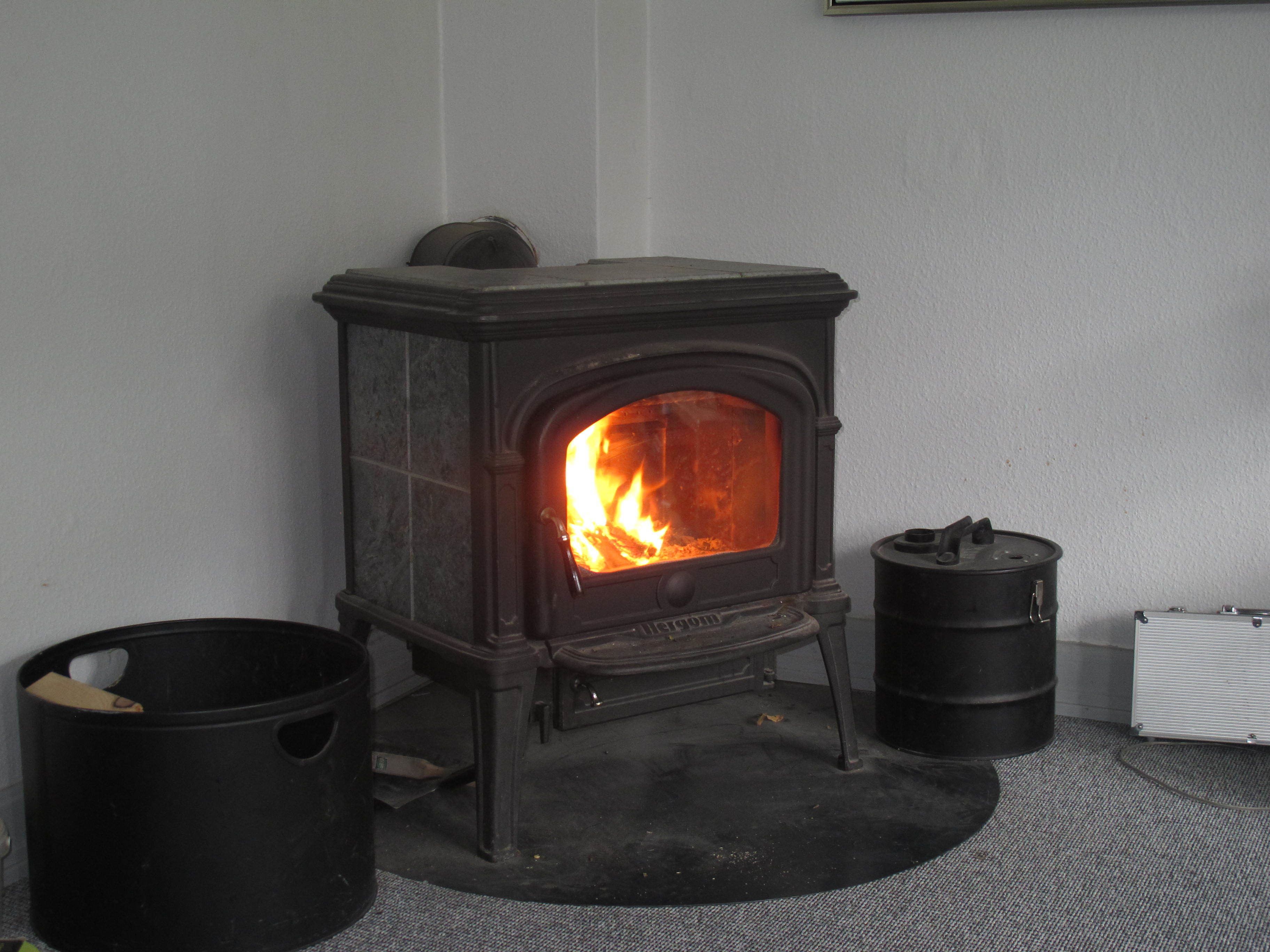 Una estufa de leña con fuego a la vista, un cubo de basura negro y un radiador en el suelo.