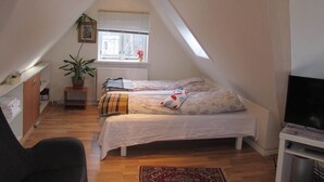 A small, cozy attic bedroom with a sloped ceiling, a bed with white bedding, a small table, a chair, a radiator, and a window with blinds. - Guesthouse Sharon Aarhus (Aarhus)