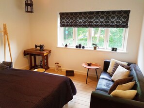 A cozy room with a bed, a wooden table with a sewing machine, a small table with a bowl, a sofa with cushions, and a window with a view of greenery. - Køge Guesthouse (Koge)