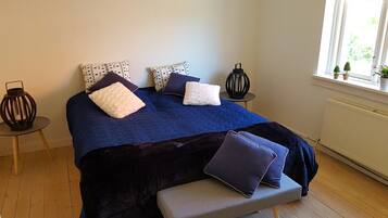 Un dormitorio con cama, un banco, una mesita con un farol y una ventana con plantas.