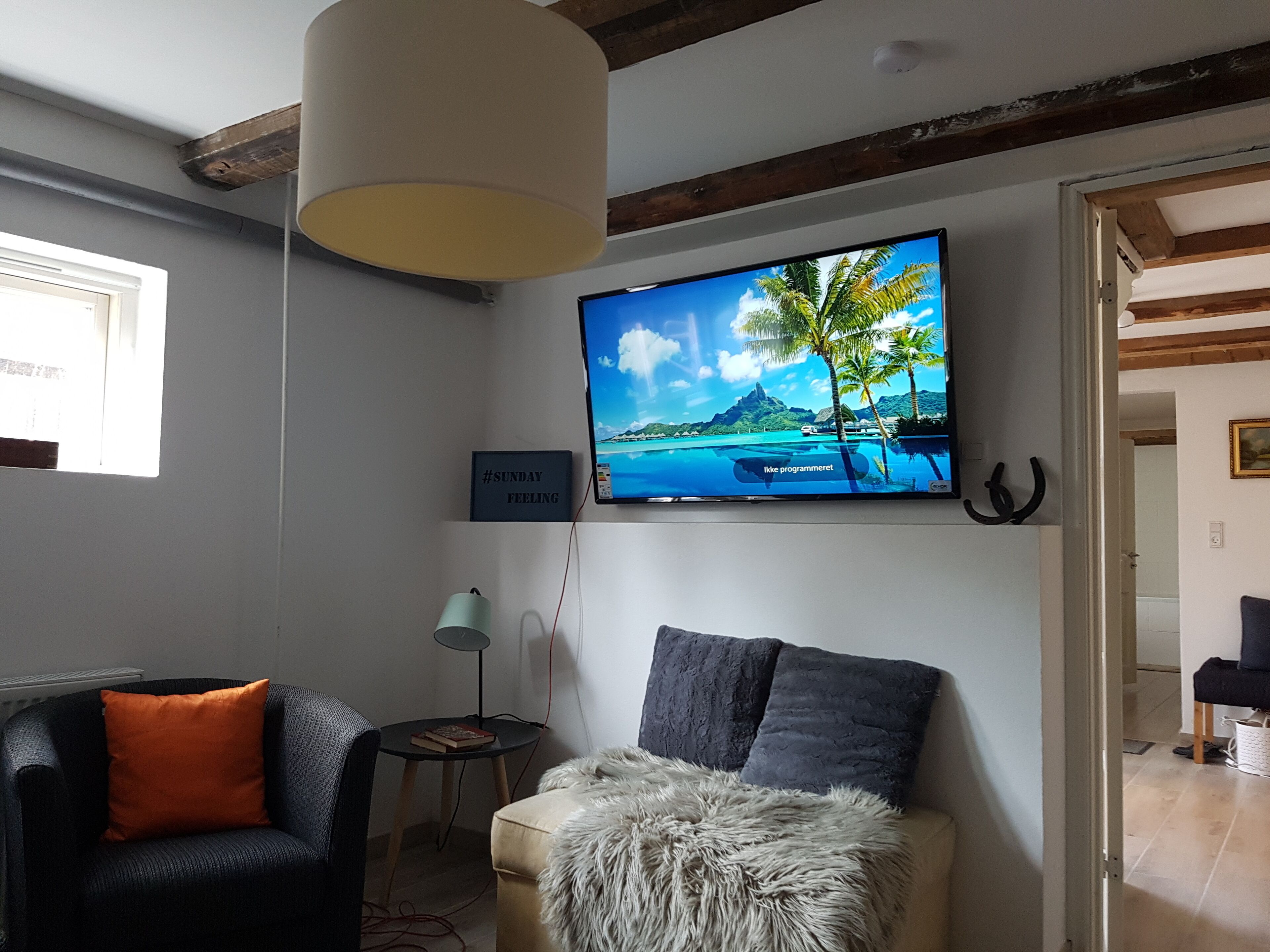 A living room with a television mounted on the wall displaying a tropical beach scene, a sofa with a fluffy throw, a small side table, and a lamp.