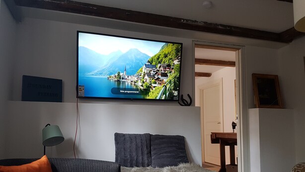A living room with a television displaying a scenic landscape, a sofa, a lamp, and a door leading to another room. - Køge Guesthouse (Koge)