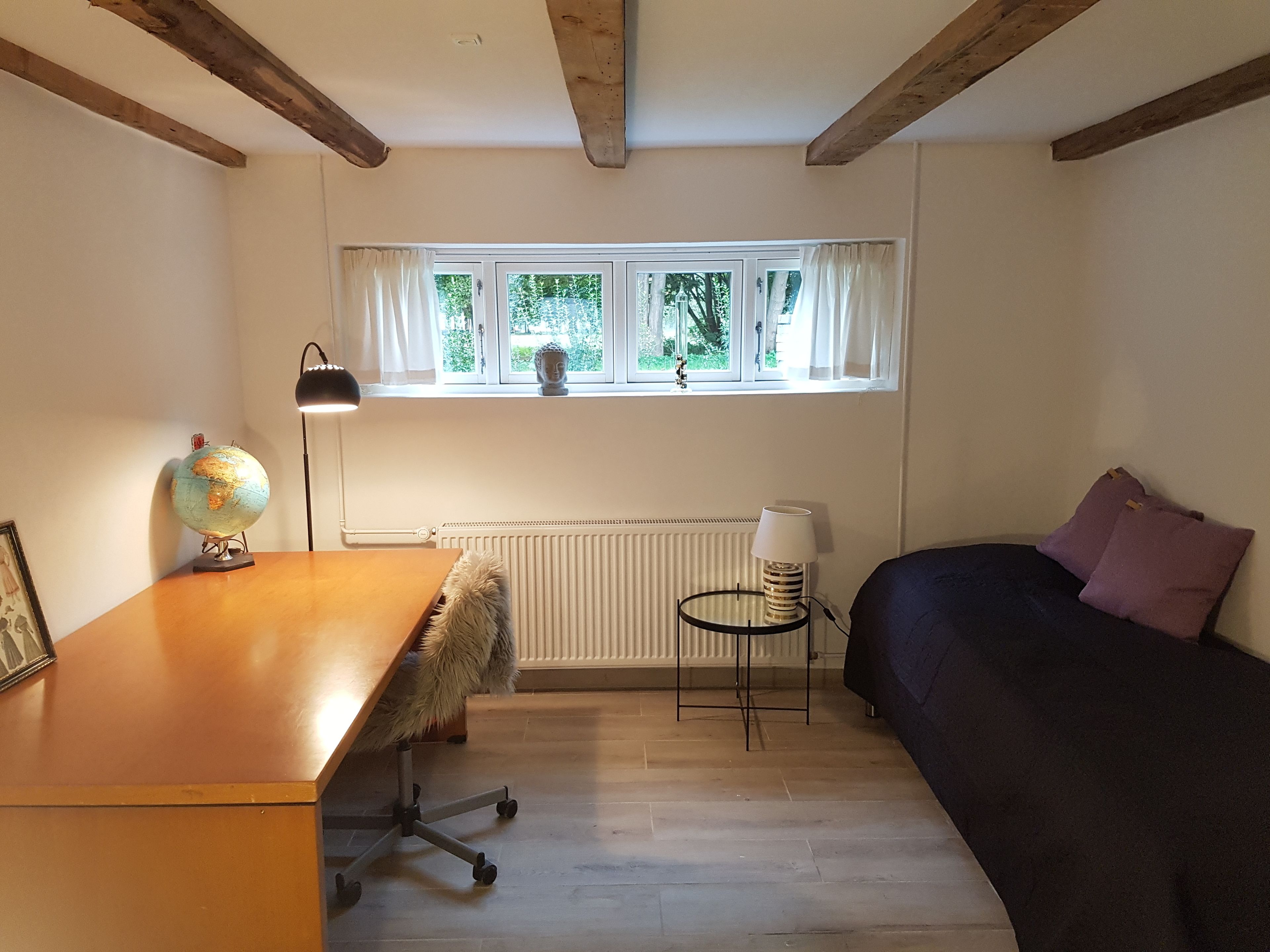 A room with a wooden desk, a globe, a lamp, a radiator, a window with a view of greenery, a bed with a dark cover, and a small table with a lamp.
