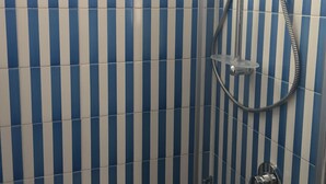 A shower with a hose and a handheld showerhead, set against a wall with blue and white vertical stripes. - Sole Flat (Minori)