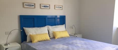 A bedroom with a blue headboard, two bedside tables, a bed with a striped bedspread, and three framed fish wall art pieces.