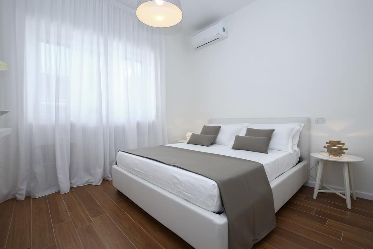 A modern bedroom with a bed, curtains, an air conditioning unit, and a small table with a lamp.