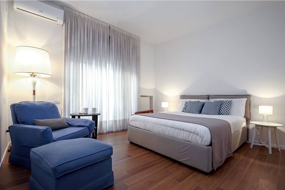 A modern hotel room with a large bed, a blue armchair, a small table, and a standing lamp.