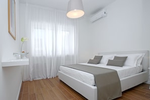 Un dormitorio moderno con una cama, cortinas blancas, una mesita de noche con un florero y un lámpara colgante.