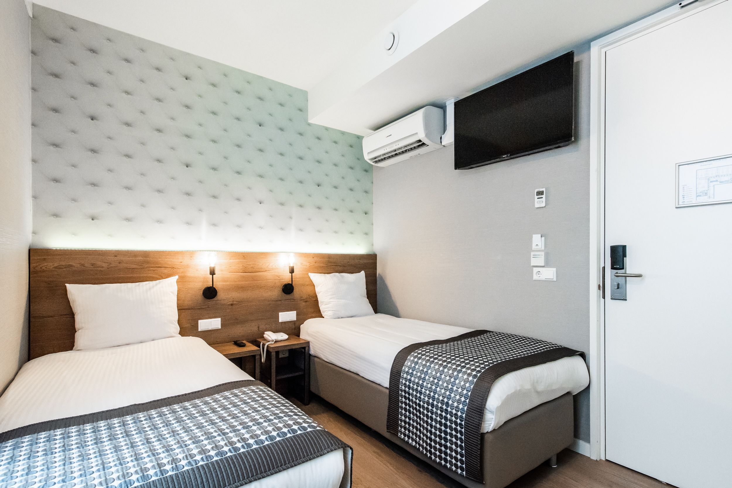 A hotel room with two beds, a wooden headboard, a wall-mounted TV, and an air conditioning unit.