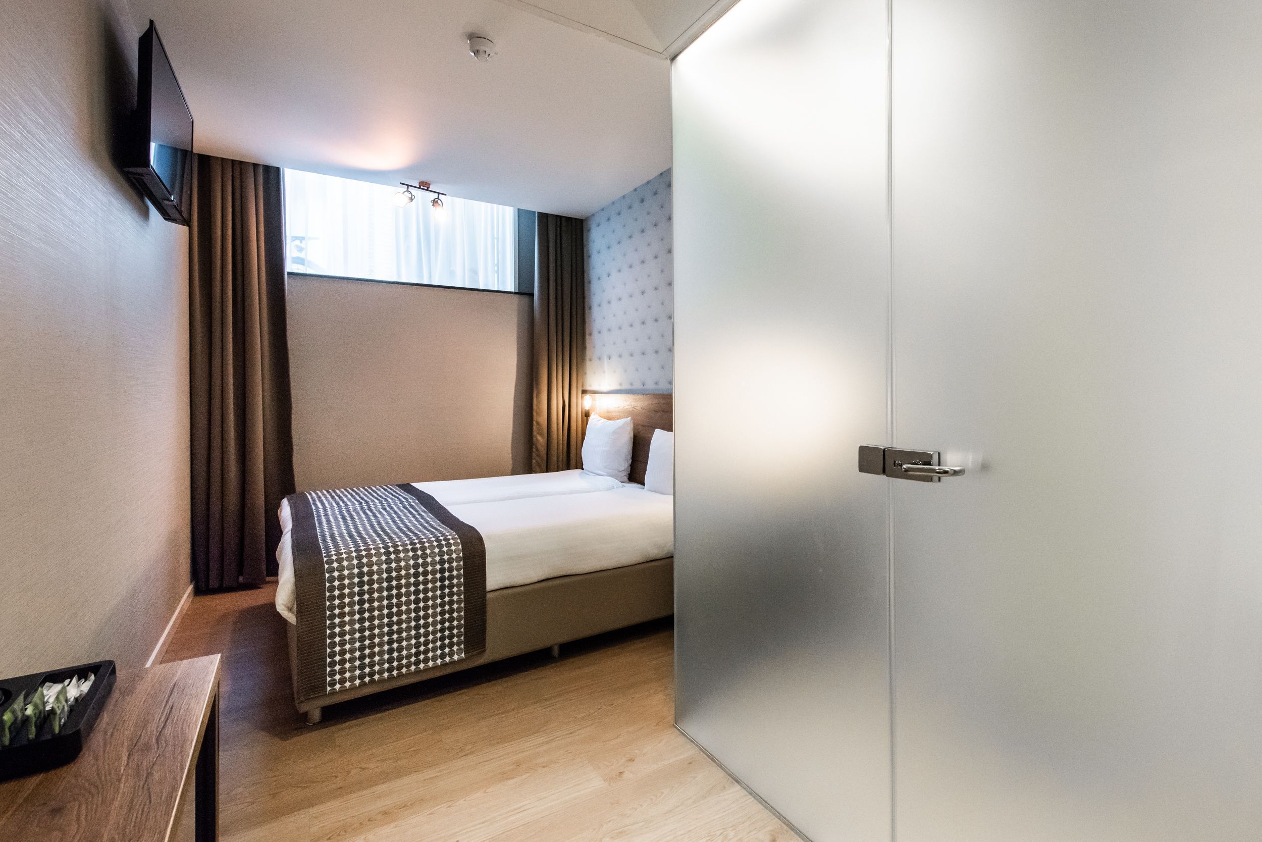 A hotel room with a bed, a bedside table, a TV mounted on the wall, and a door.