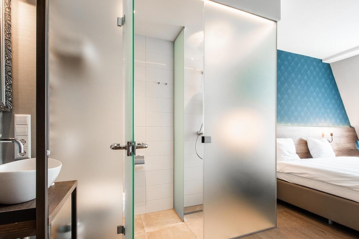 A modern hotel room with a glass partition separating the bathroom and bedroom.