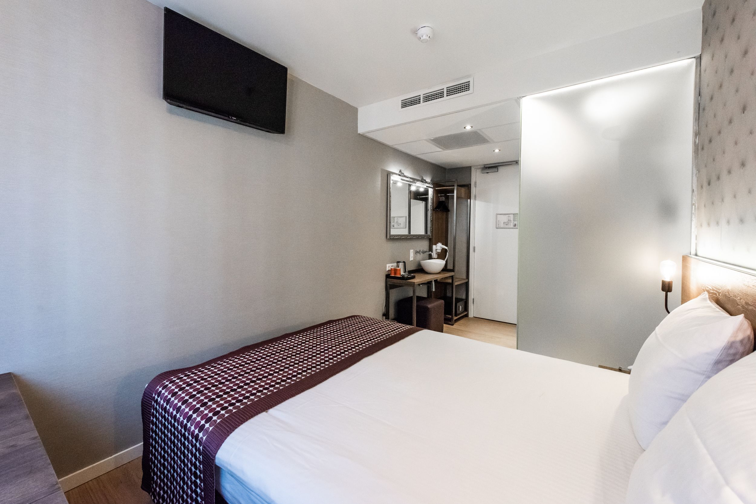 A modern hotel room with a large bed, a bedside table, a mirror, and a wardrobe.