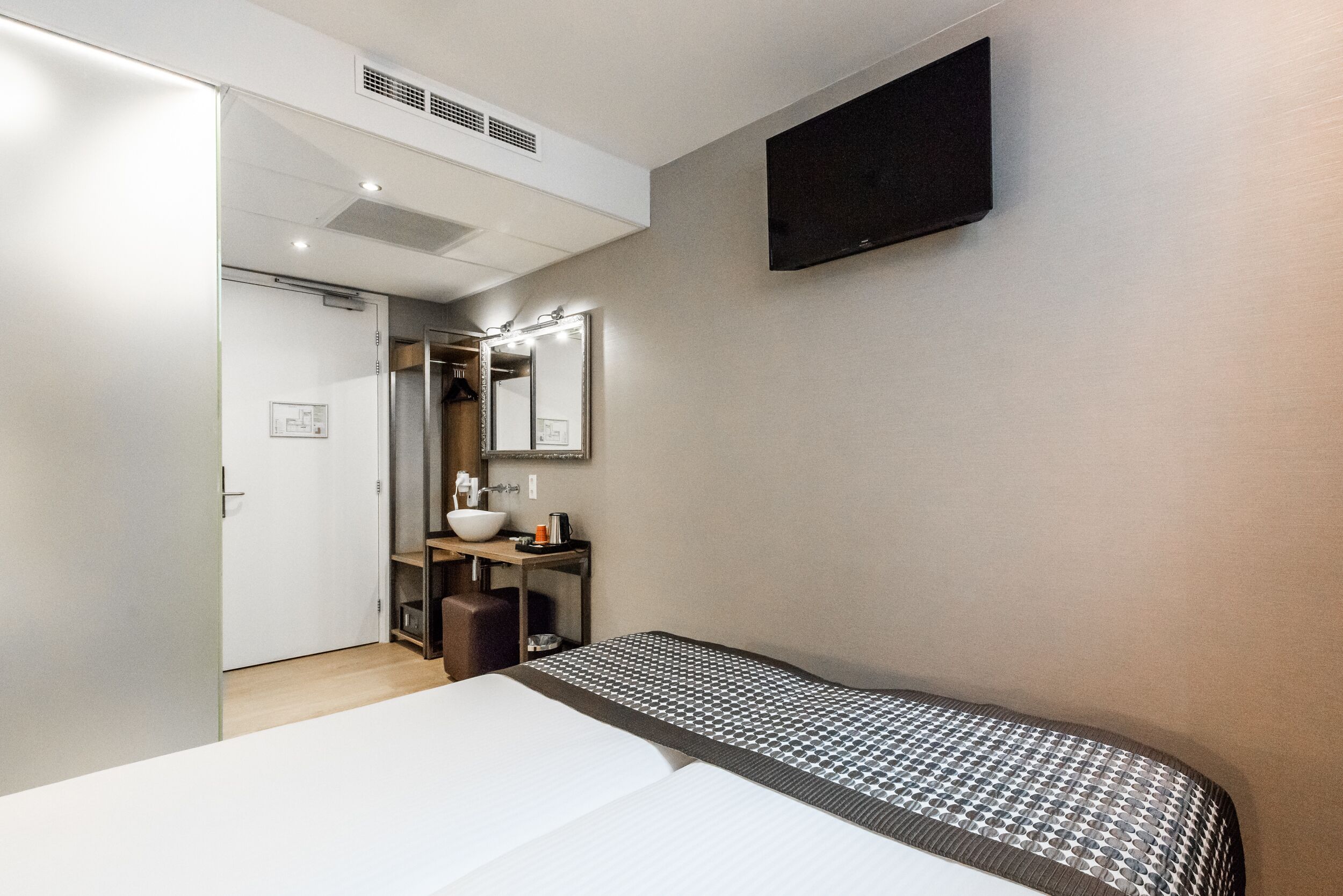 Een hotelkamer met een bed, nachtkastje, spiegel, tv aan de muur en een badkamer met wastafel en spiegel.